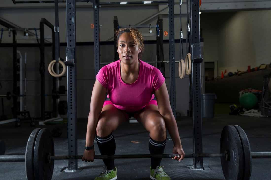 A young Black woman in a pink shirt at a gym hovering over a barbell in a deadlift position.