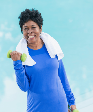 Smiling older Black woman in a blue shirt with a towel around her neck holding green dumbells.