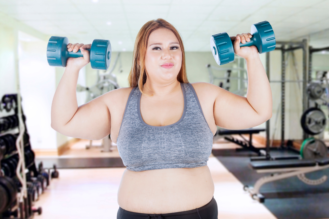 Smiling woman in a gym lifting dumbbells above her shoulders.